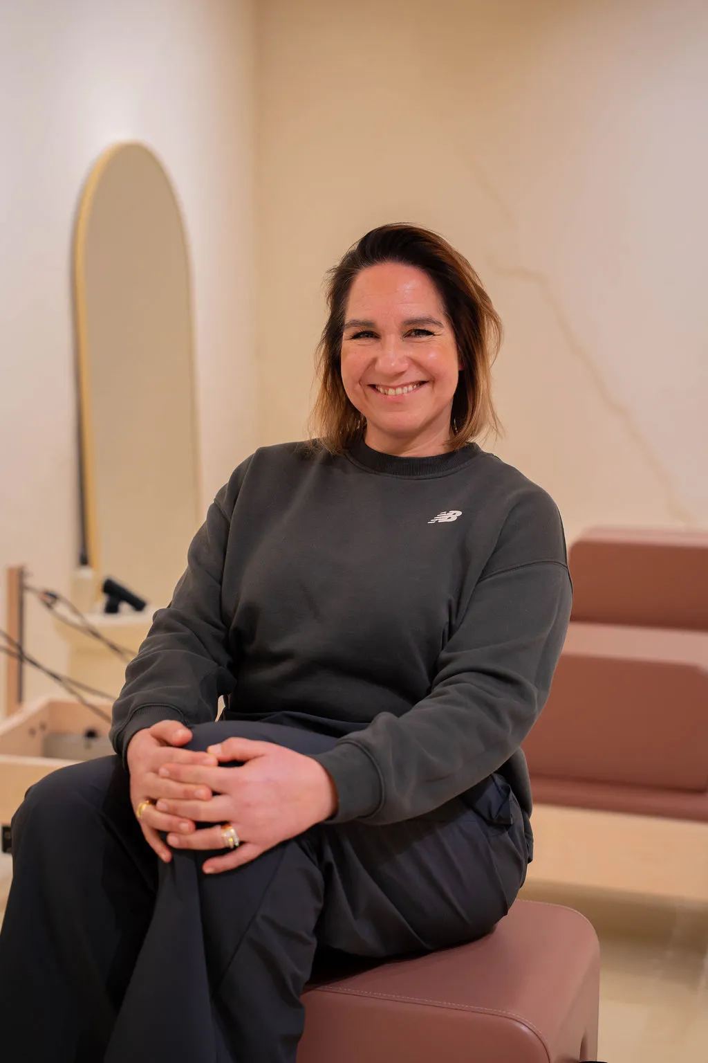 Een lachende vrouw met schouderlang haar, gekleed in een donkere sweater, zit op een gestoffeerde bank in een lichtgekleurde kamer met een grote spiegel op de achtergrond.