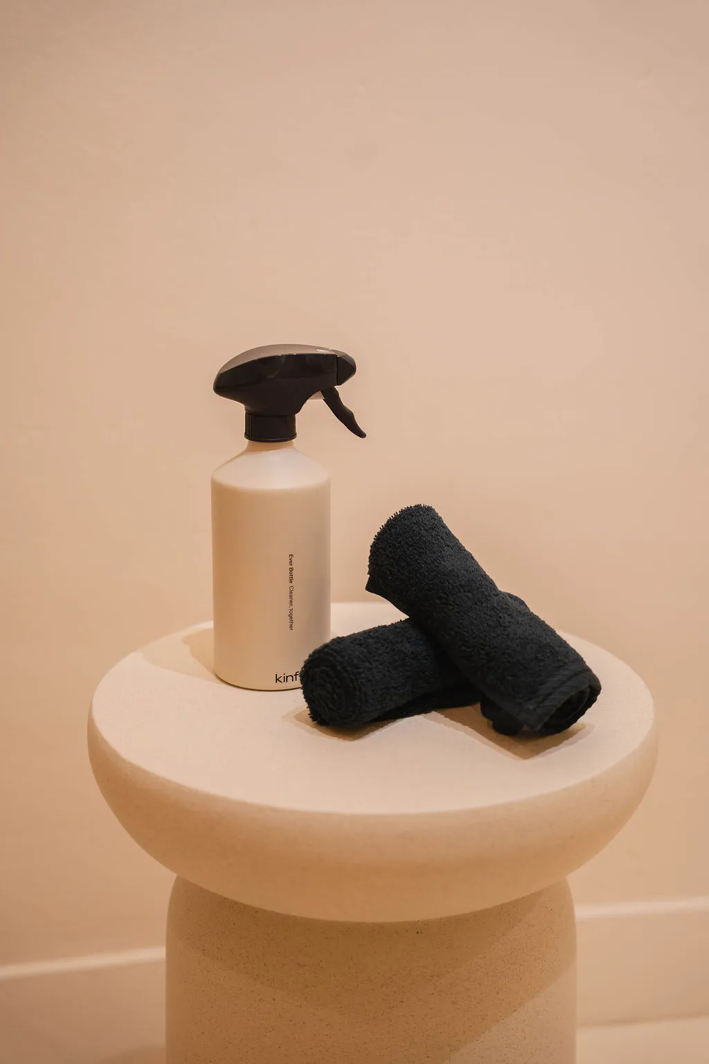 Beige spuitfles met zwarte trekker en twee opgerolde zwarte handdoeken op een ronde beige sokkel tegen een beige achtergrond.
