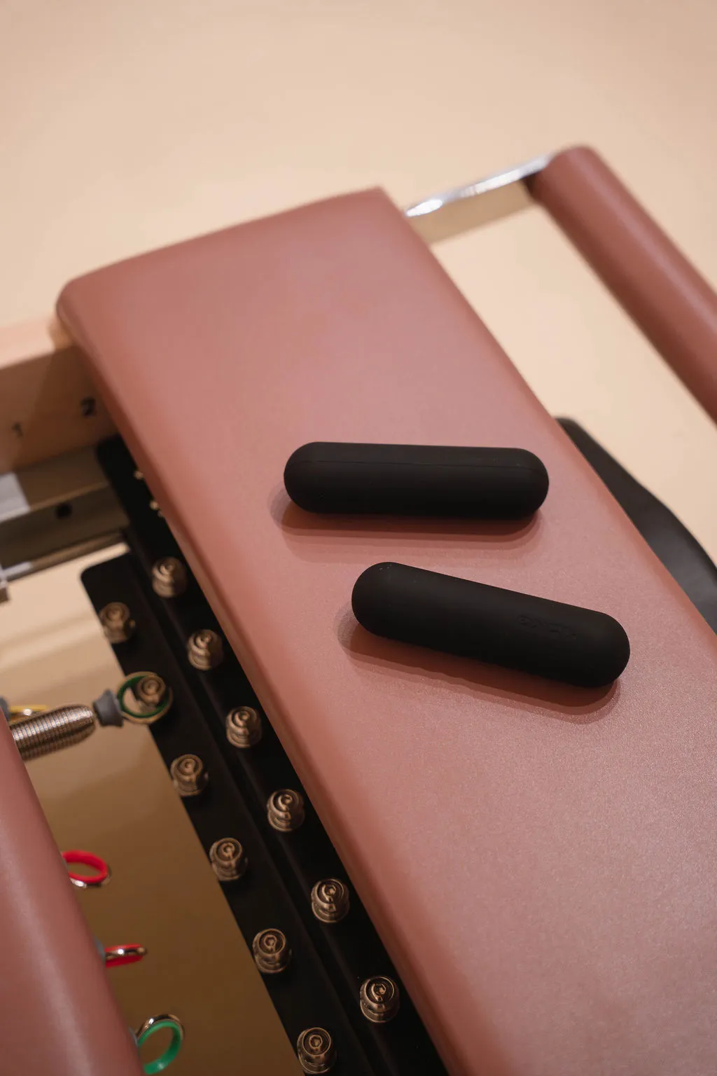 Detailopname van twee zwarte cilindrische schuimrubberen handgewichten die rusten op een roze, gewatteerd oppervlak van een reformer met metalen verstelknoppen.