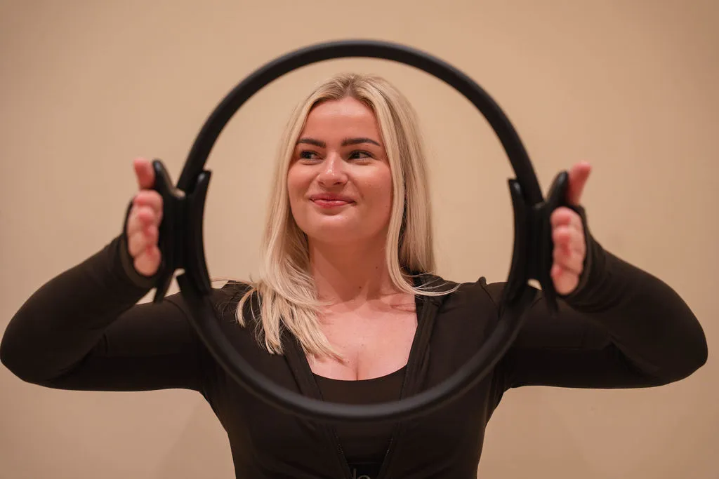 Lachende vrouw met opgestoken haar die een zwarte fitnessring naar de camera richt.