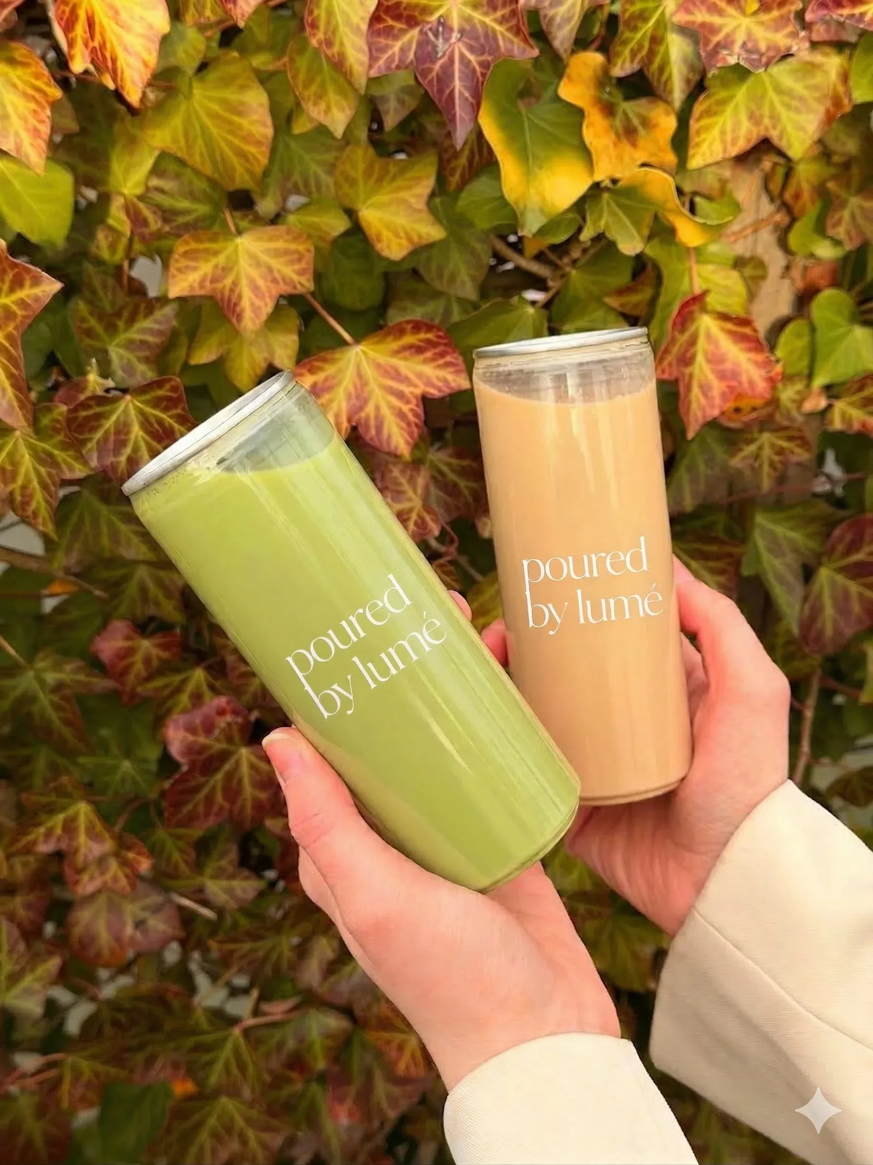 Handen houden twee blikjes drank vast met het opschrift 'poured by Lumé', een met een groene en een met een beige drank, tegen een achtergrond van groene en roodachtige bladeren.