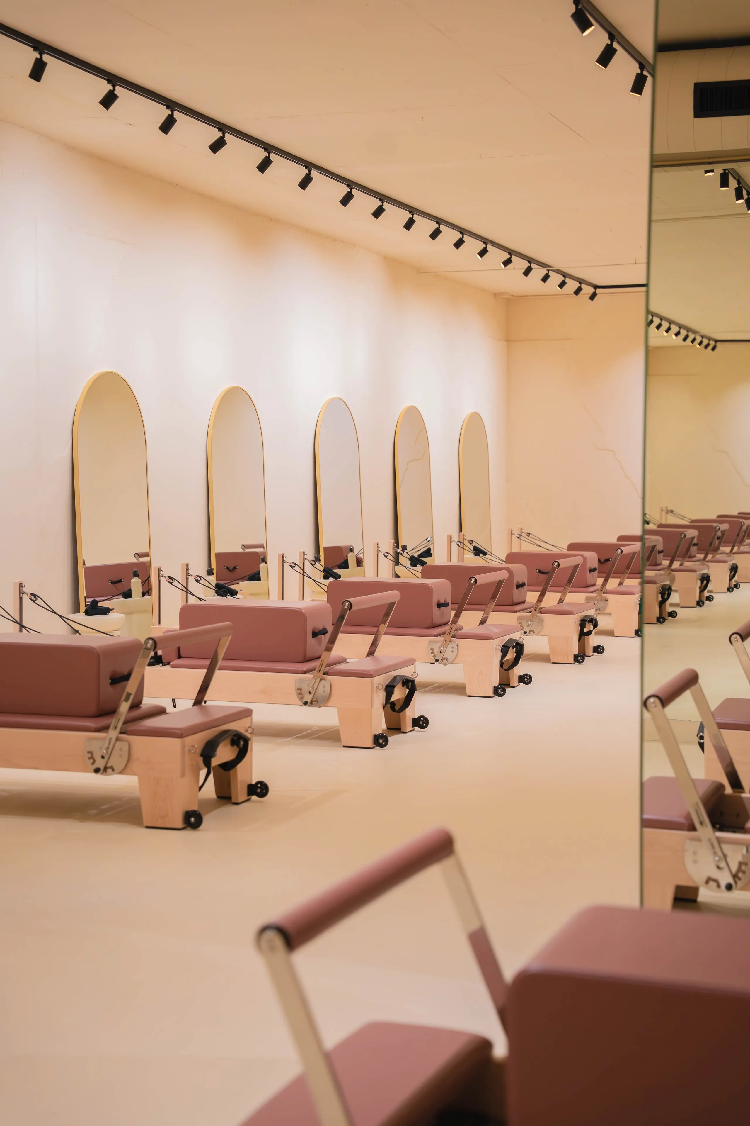 Een rij Pilates reformer-apparaten met houten frames en roze bekleding in een minimalistische studio met gebogen spiegels en railverlichting.