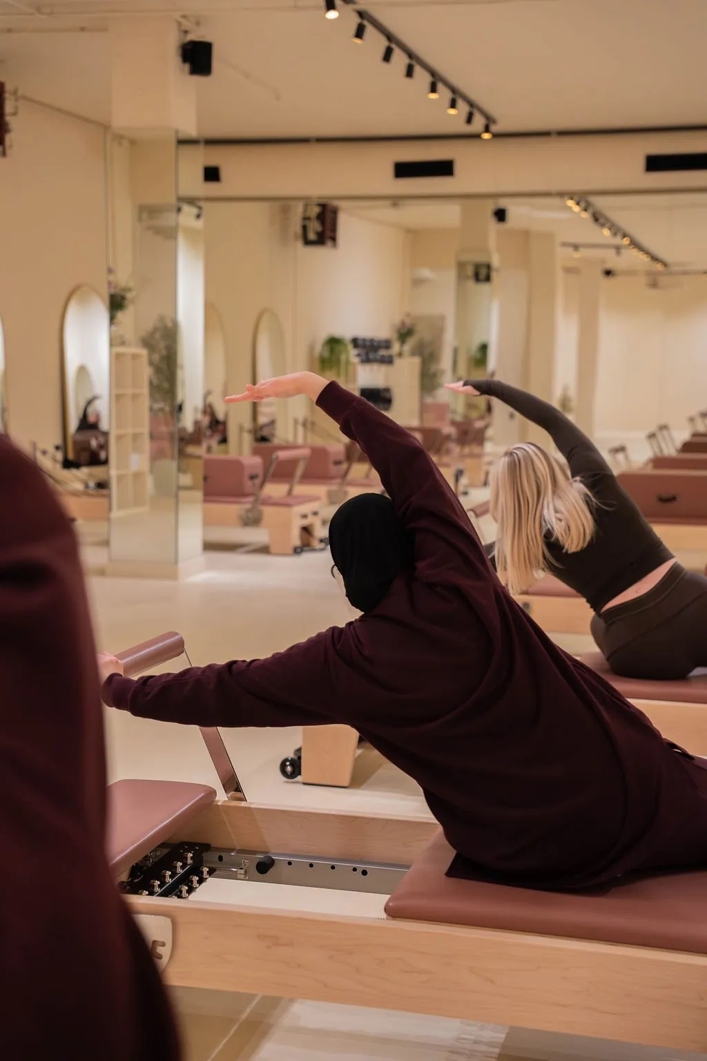 Twee vrouwen doen zijwaartse rekoefeningen op Pilates reformer-apparaten in een lichte fitnessstudio met spiegels.