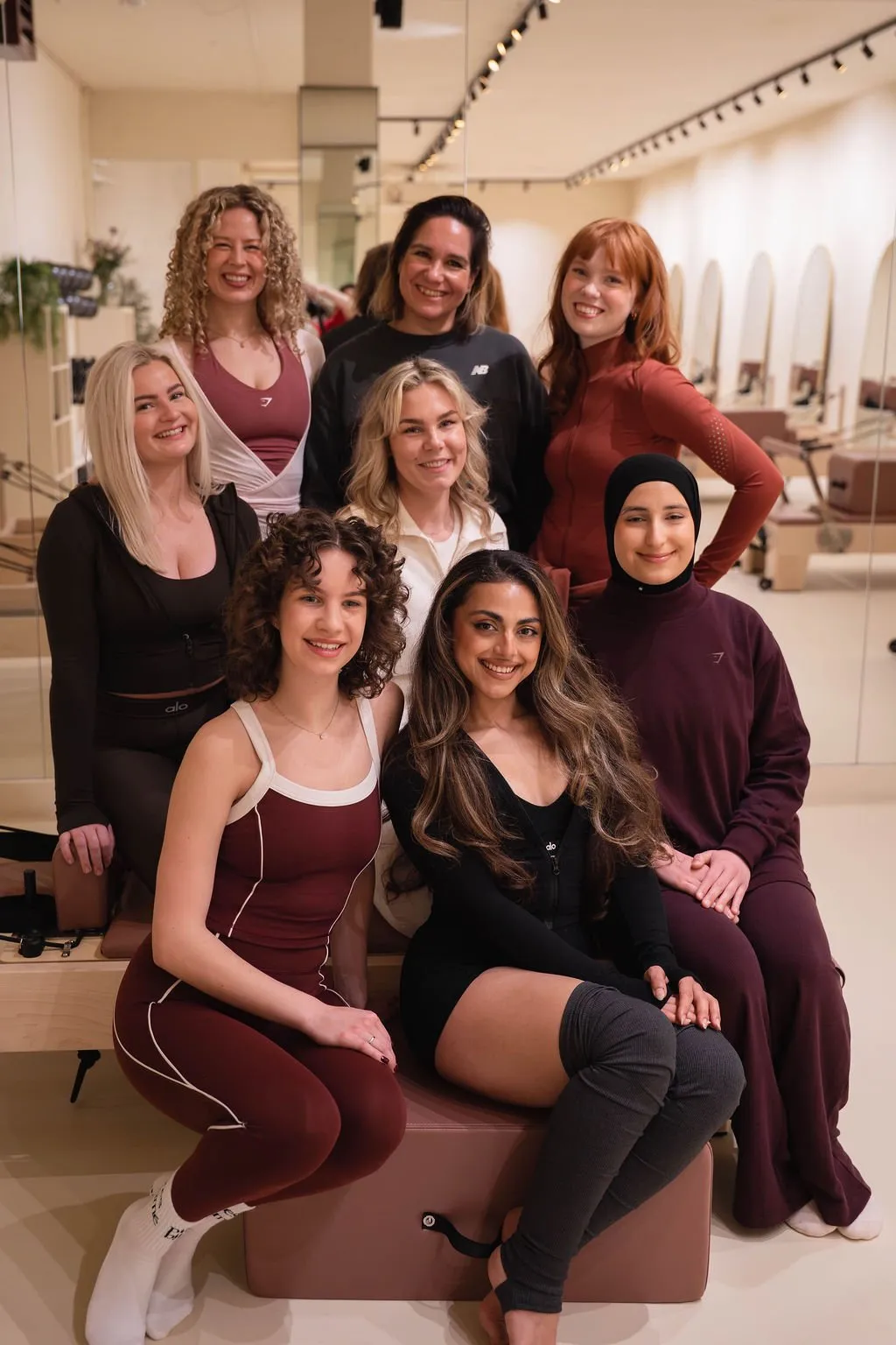 Een groep van acht diverse vrouwen lacht en poseert samen in een lichte ruimte met spiegels en pilatesapparatuur. Ze dragen sportkleding, waaronder leggings en topjes.