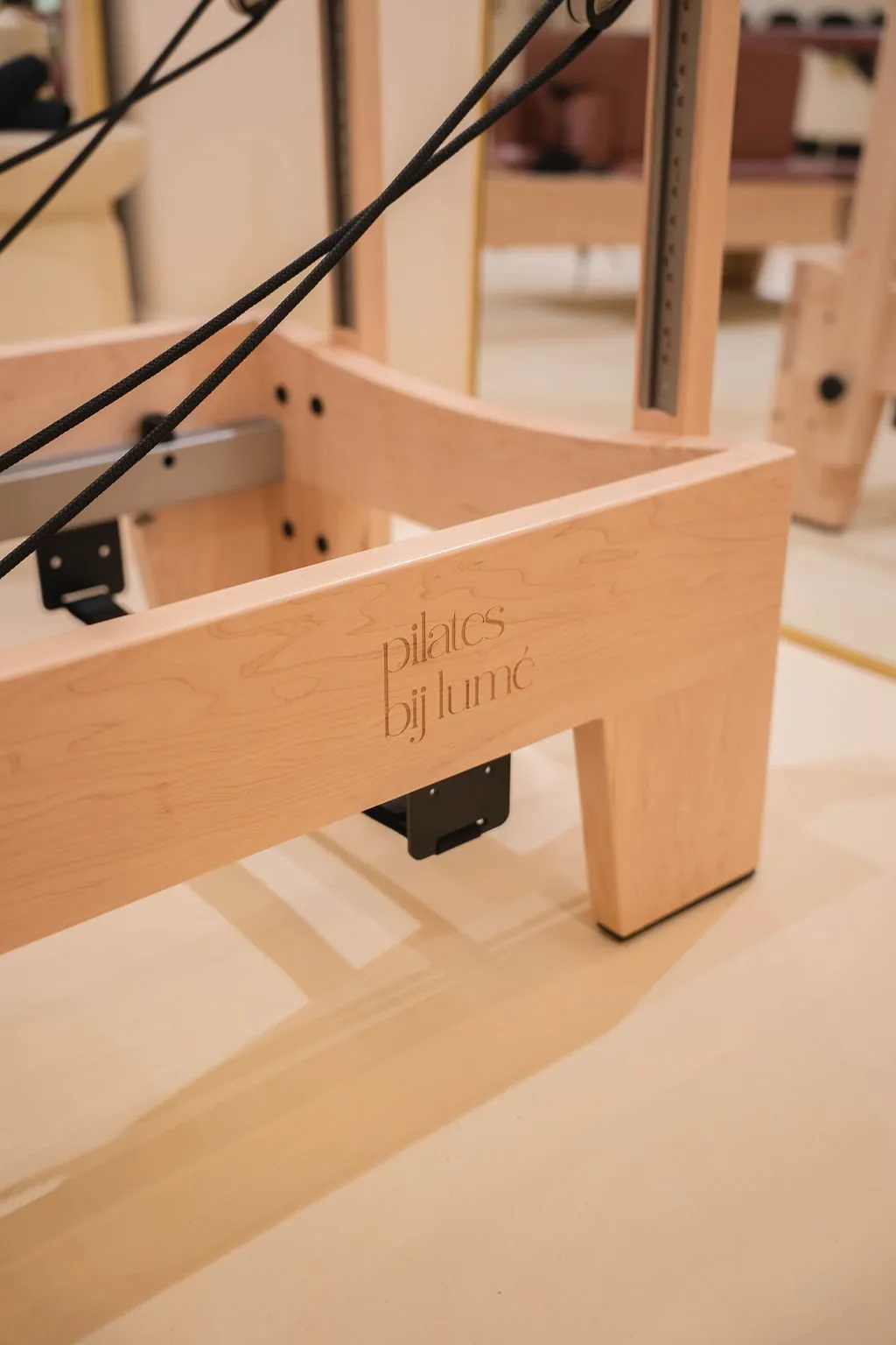 Detailopname van een licht houten frame van een Pilates reformer machine met zwarte weerstandskoorden en de gegraveerde tekst 'pilates bij lumé' aan de zijkant.