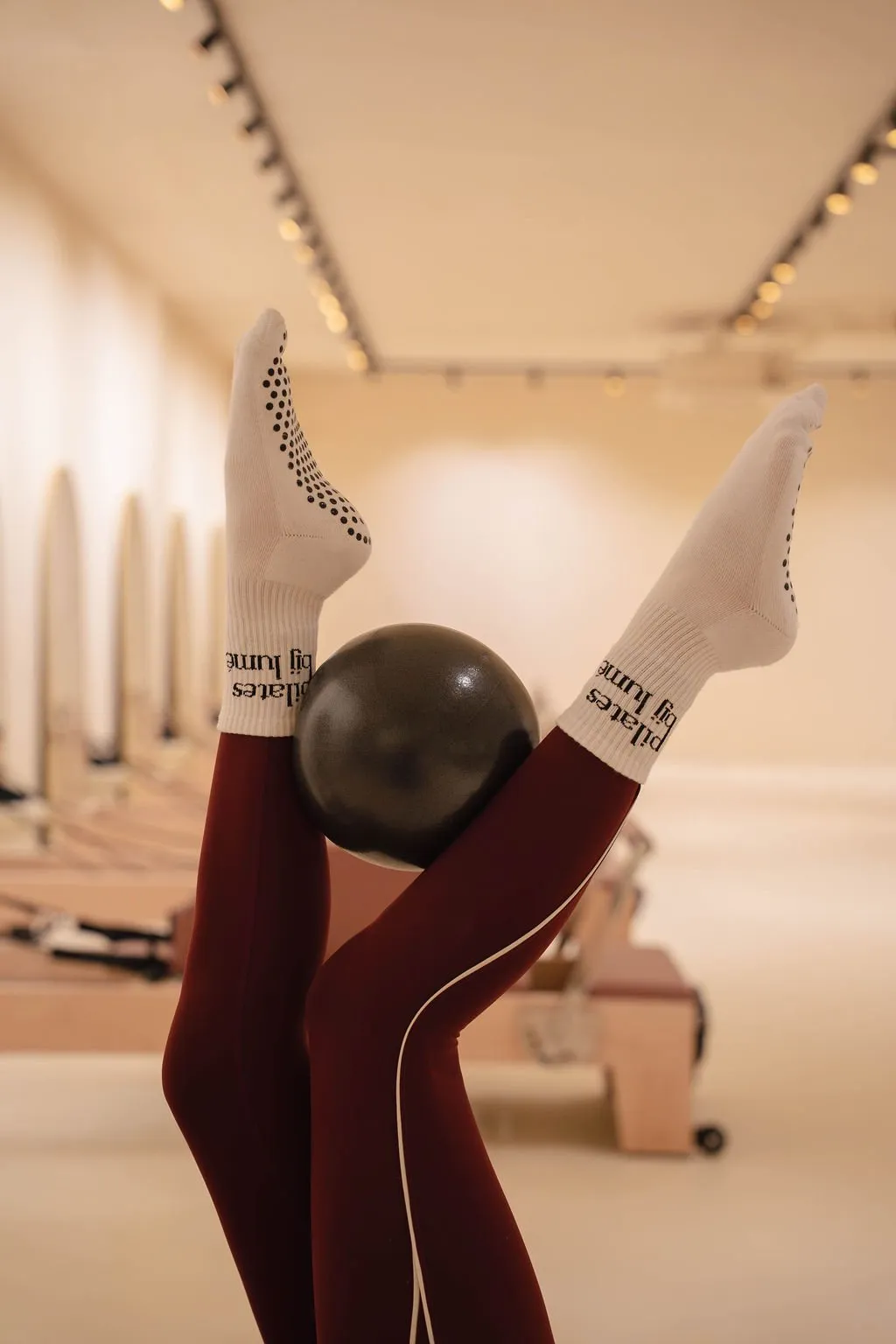 Een persoon ligt op de rug met de benen omhoog, draagt witte sokken met zwarte stippen en een bordeauxrode legging, en houdt een zwarte oefenbal tussen de voeten in een lichte pilatesstudio met houten reformers.