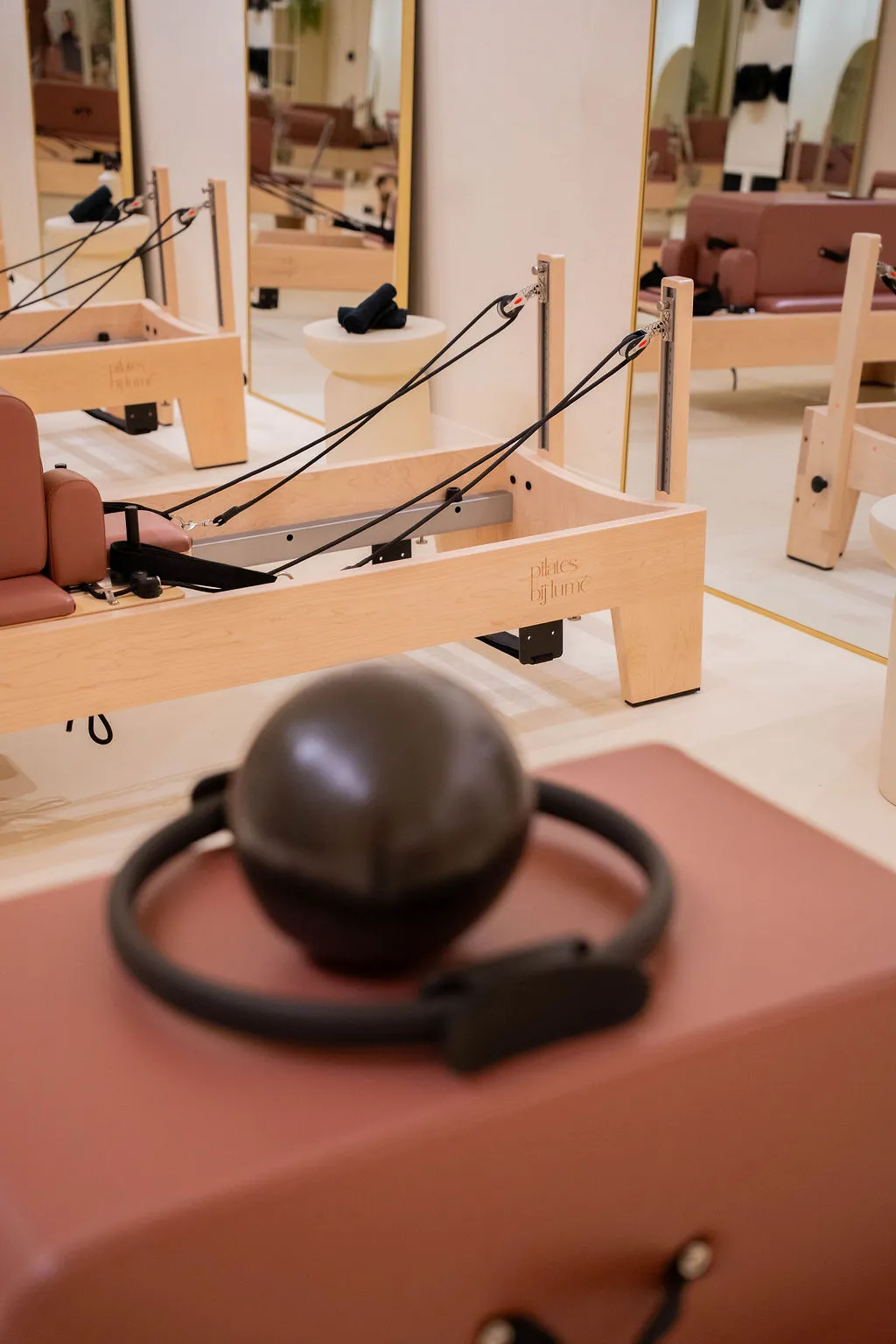 Licht houten Pilates reformer machine met zwarte bandjes en bruine demping in een lichte studio met grote spiegels, met een donkergrijze Pilates ring en bal op de voorgrond.