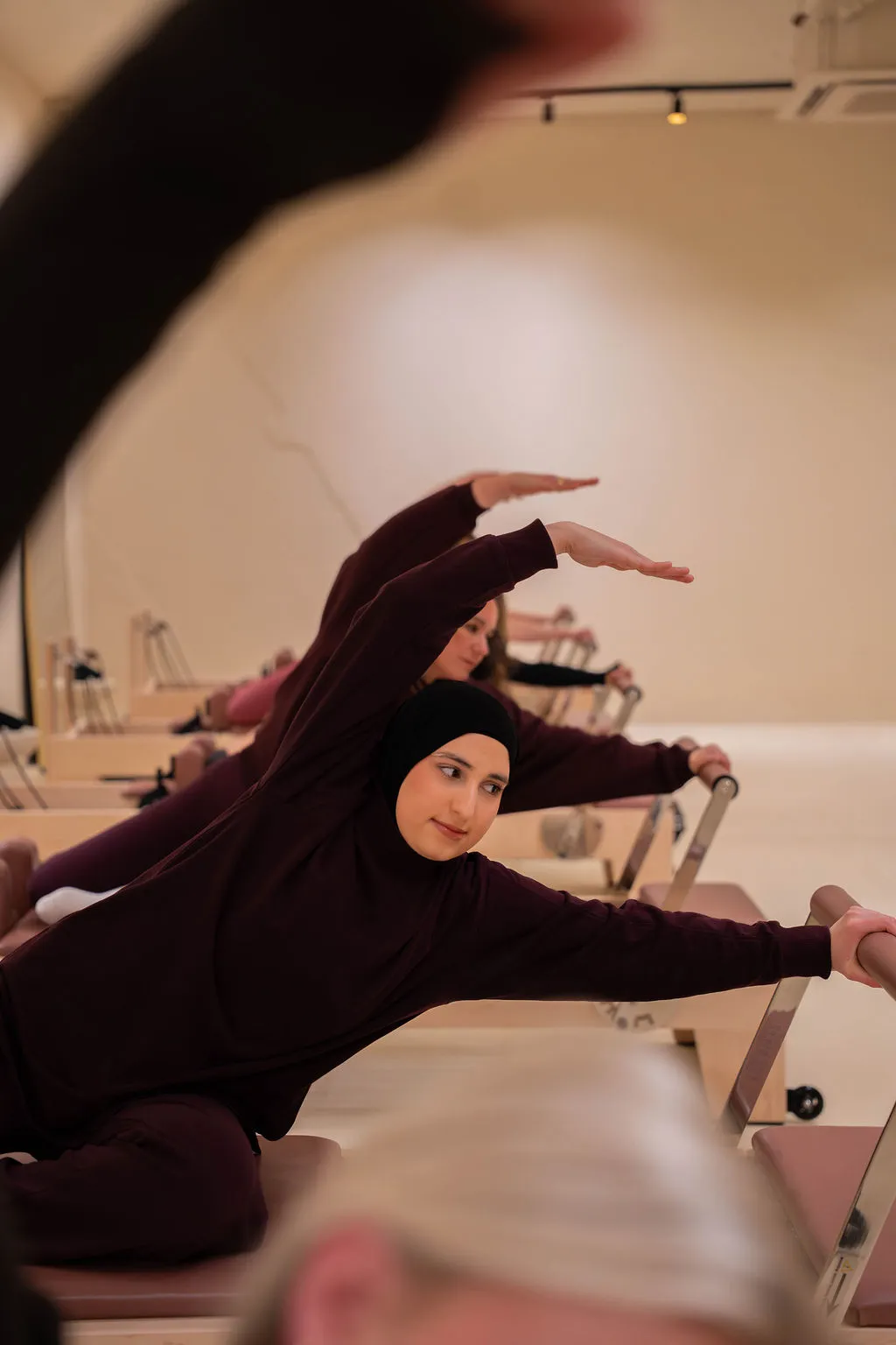Een groep vrouwen beoefent Pilates op reformer-apparaten, waarbij de aandacht uitgaat naar een vrouw met een zwarte hijab en bordeauxrode kleding die haar arm boven haar hoofd strekt terwijl ze de handgreep van het apparaat vasthoudt.