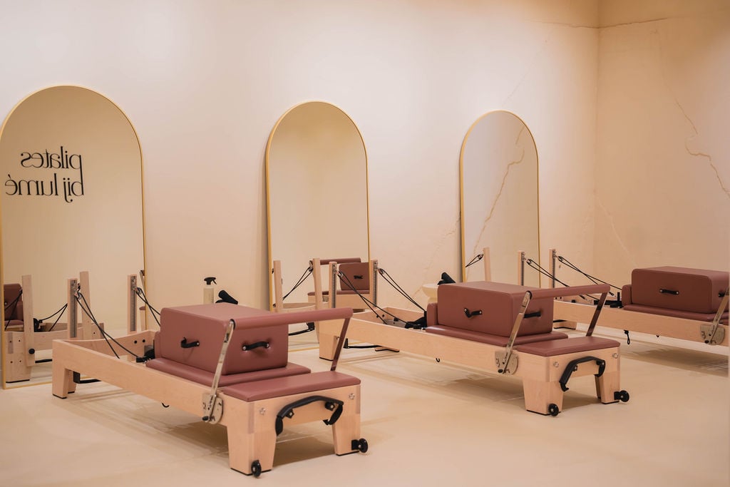 Interieur van een Pilates-studio met drie houten reformermachines, gepositioneerd voor drie hoge gebogen spiegels tegen een beige muur.