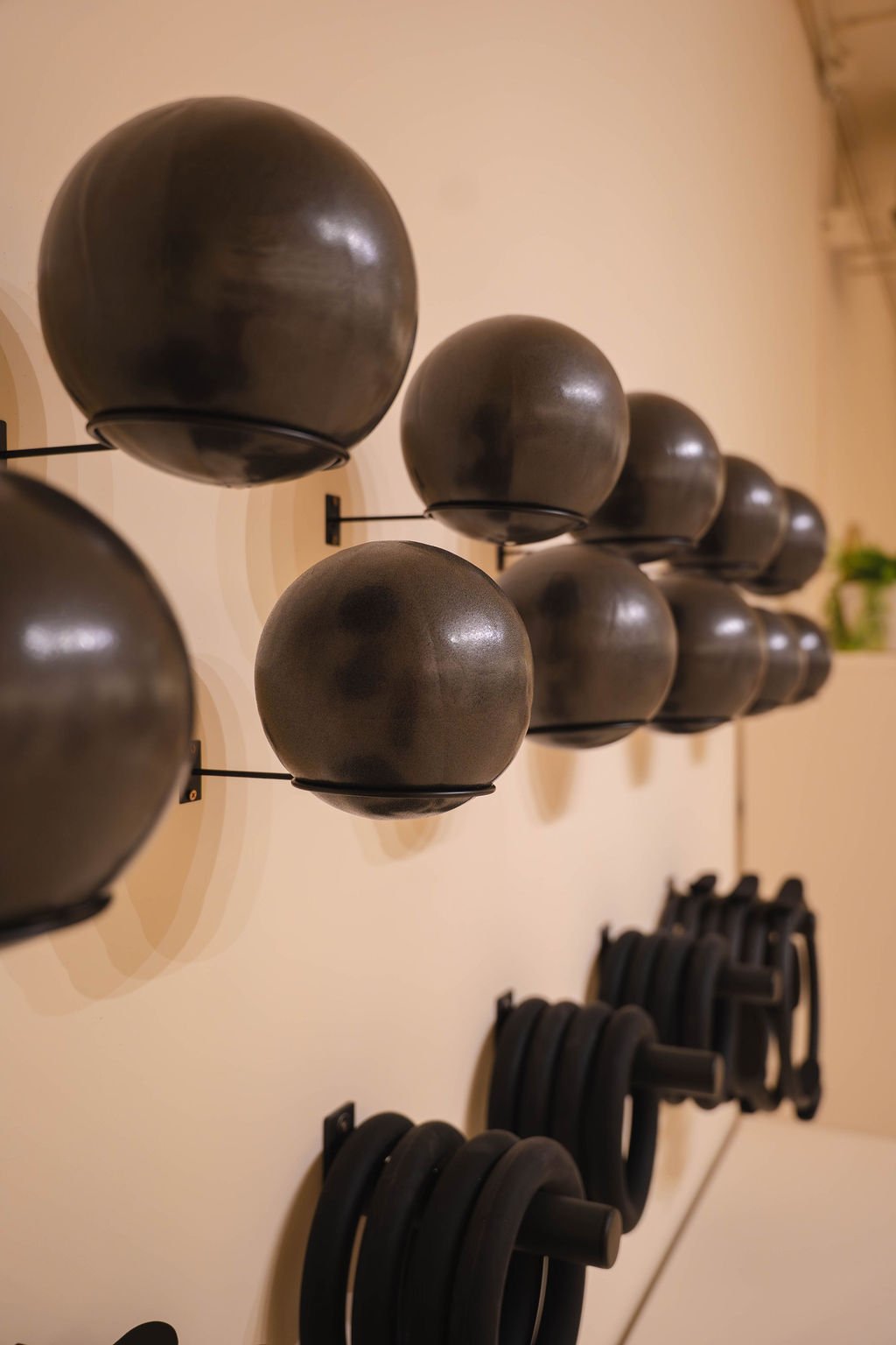 Rij zwarte medicijnballen gemonteerd op muurbeugels boven zwarte opgerolde weerstandsbanden die aan een beige muur hangen in een sportschool of fitnessstudio.