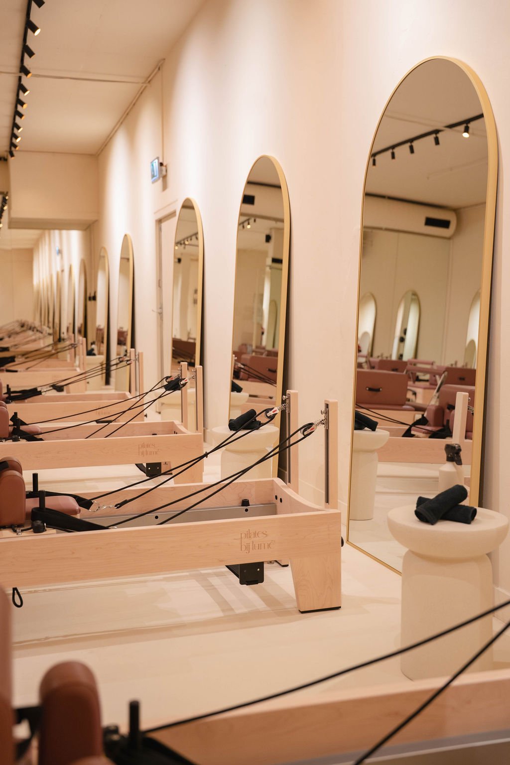 Interieur van een Pilates-studio met houten reformermachines opgesteld langs een muur met grote, gebogen spiegels die ze weerspiegelen, en een ronde kruk met gevouwen zwarte handdoeken in de buurt.