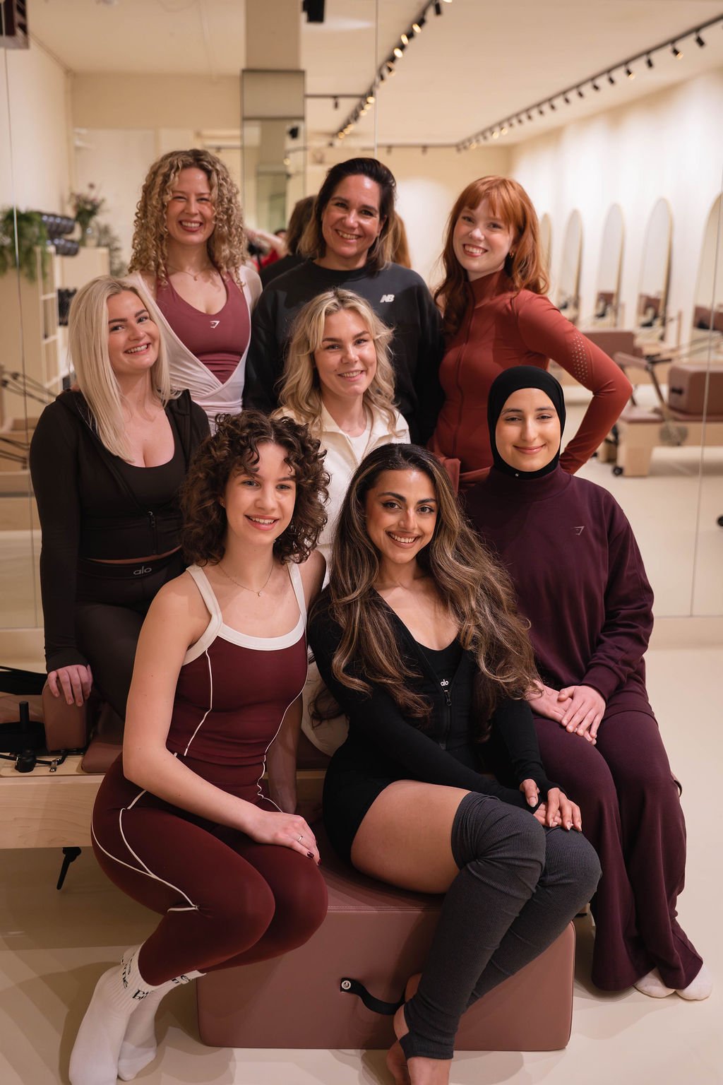 Groep van acht lachende vrouwen die samen poseren in een lichte kamer met spiegels en pilatesapparatuur, gekleed in actieve kleding in de tinten zwart, wit, bordeaux en kastanjebruin, met één vrouw die een hijab draagt.