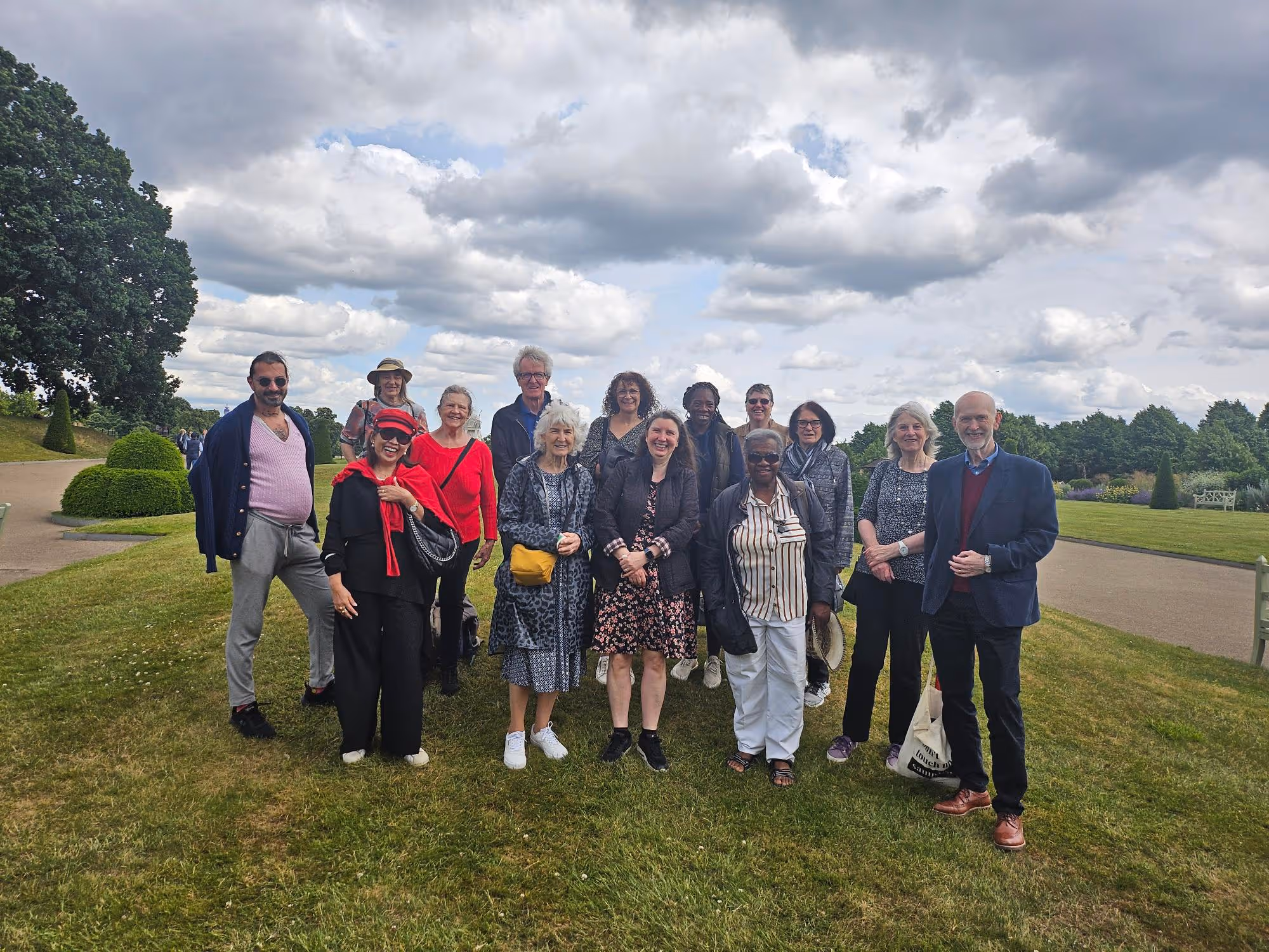 Group of fifteen diverse adults standing on grass in a park under a cloudy sky.