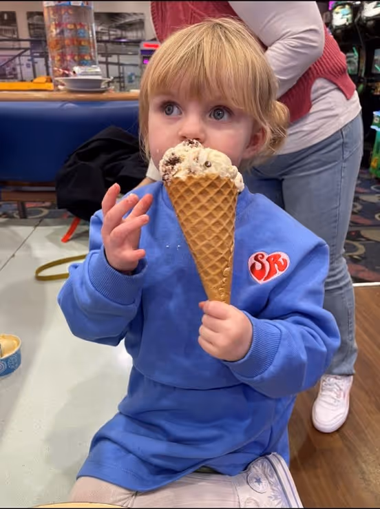 Young child in blue sweater holding and eating a large ice cream cone indoors.