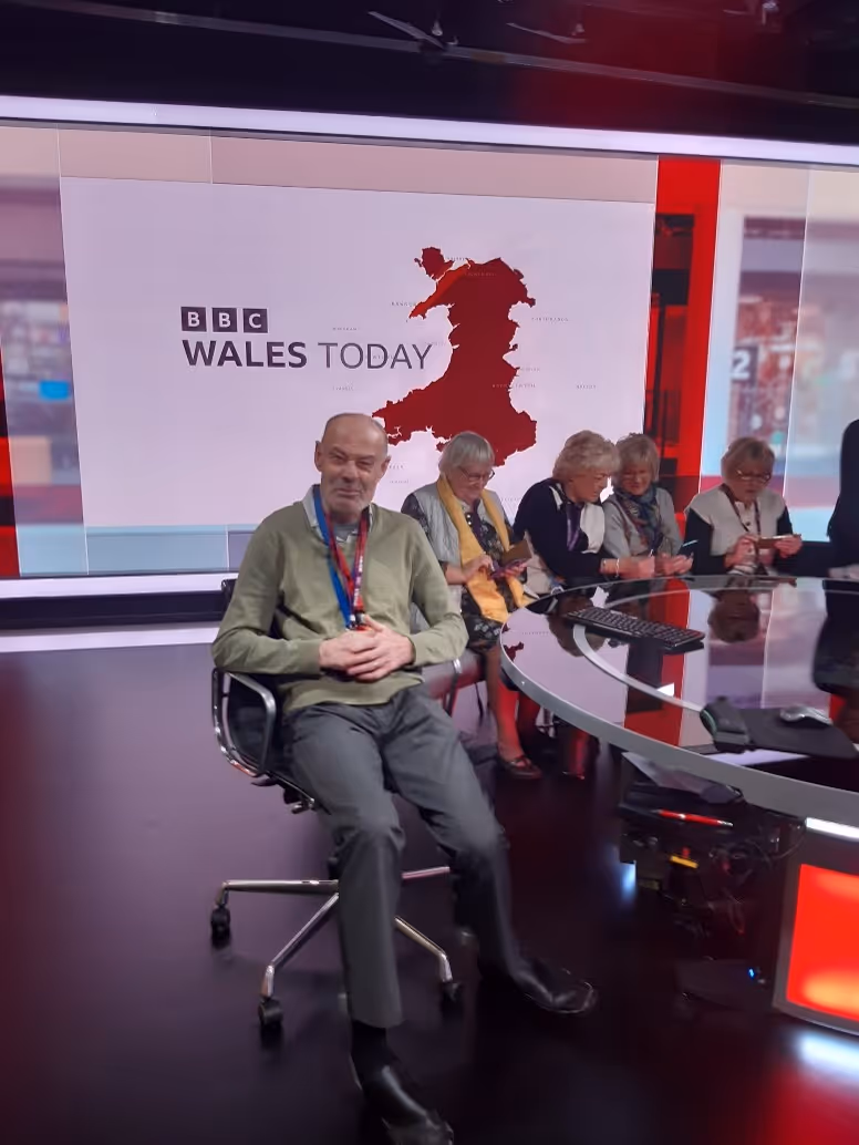 Man sitting on an office chair in a BBC Wales Today studio with four older women sitting at a round desk behind him.