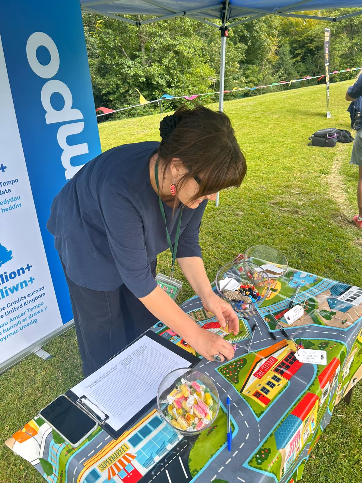 Woman with dark hair and glasses arranging items on a colorful table with a road map design under a canopy outdoors.