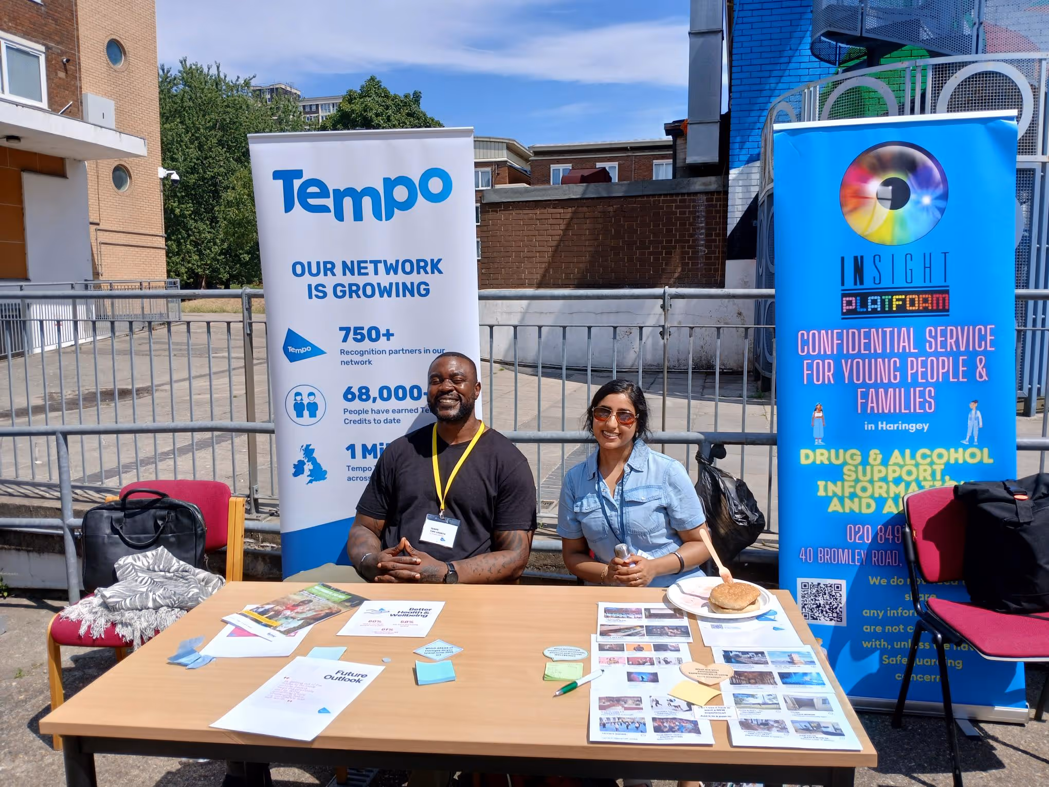 Two people sitting at an outdoor information table with banners for Tempo and Insight Platform promoting network growth and confidential youth services.