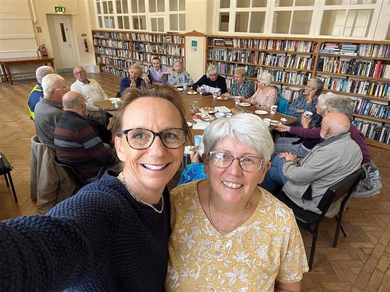 Two middle-aged women smiling for a selfie in front of a group of elderly people sitting around a table in a library room.