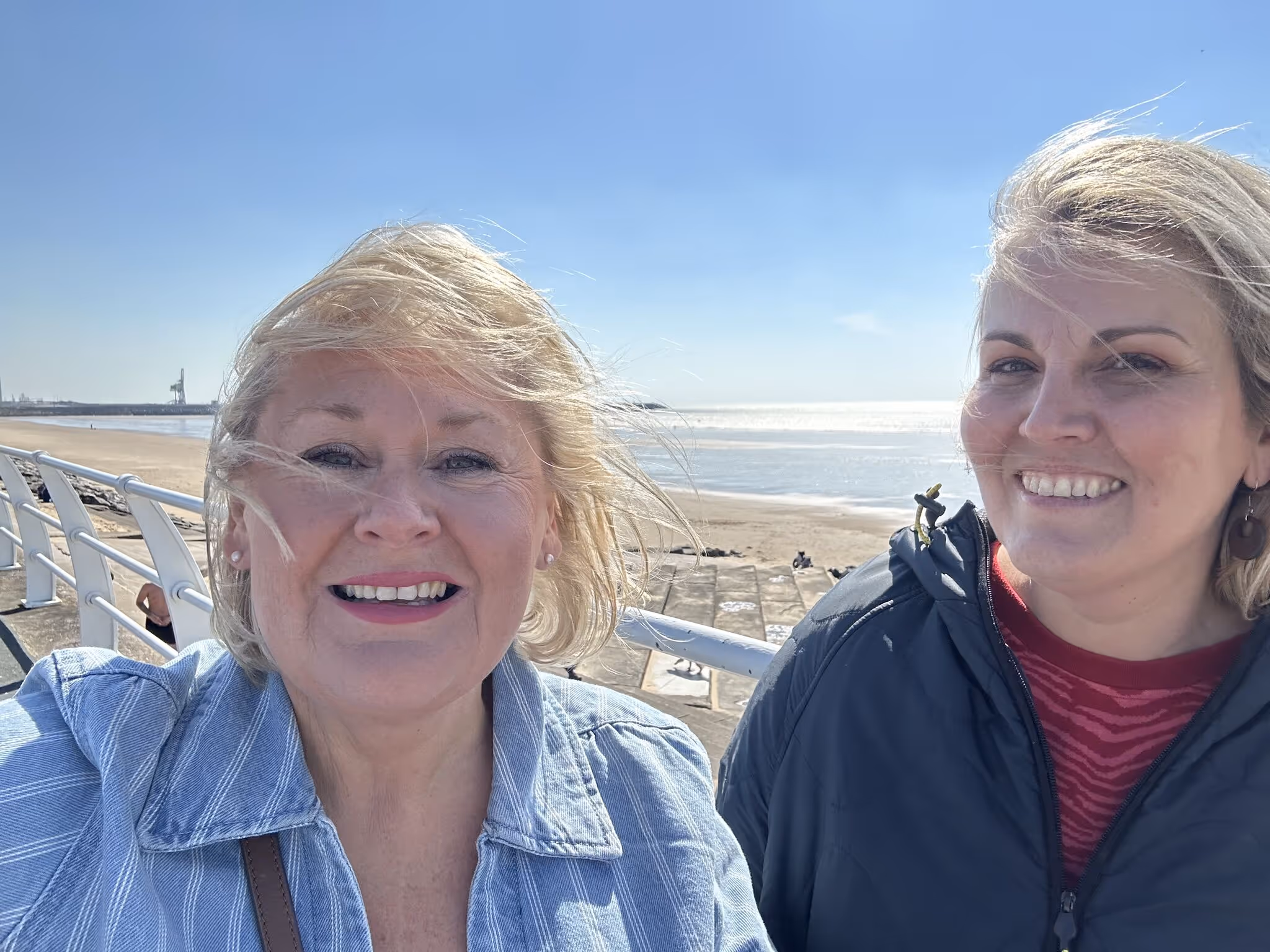 Two smiling women standing outdoors on a sunny day near a beach with clear blue sky.
