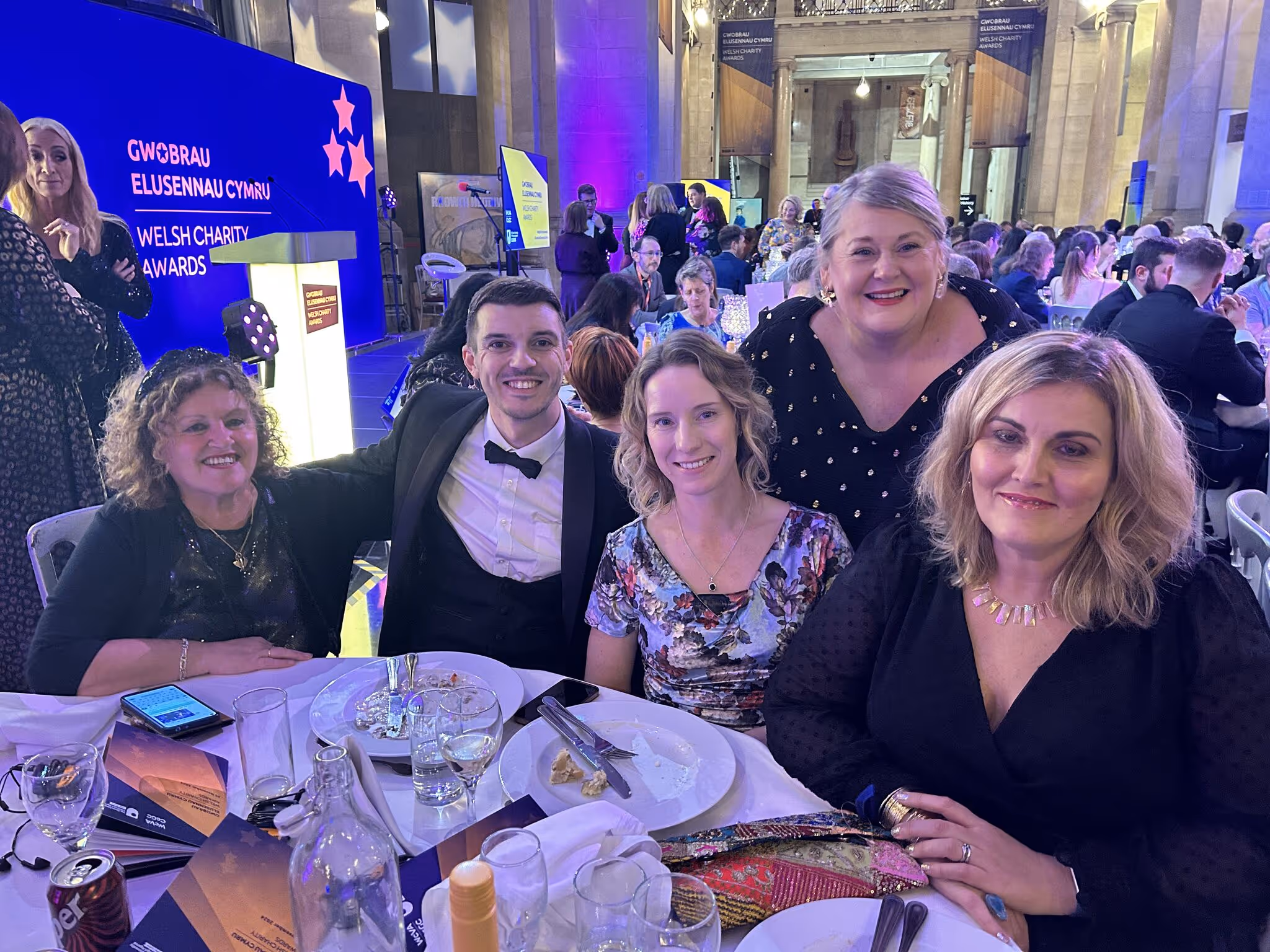 Five smiling people dressed formally seated and standing at a table during the Welsh Charity Awards event in a large decorated hall.