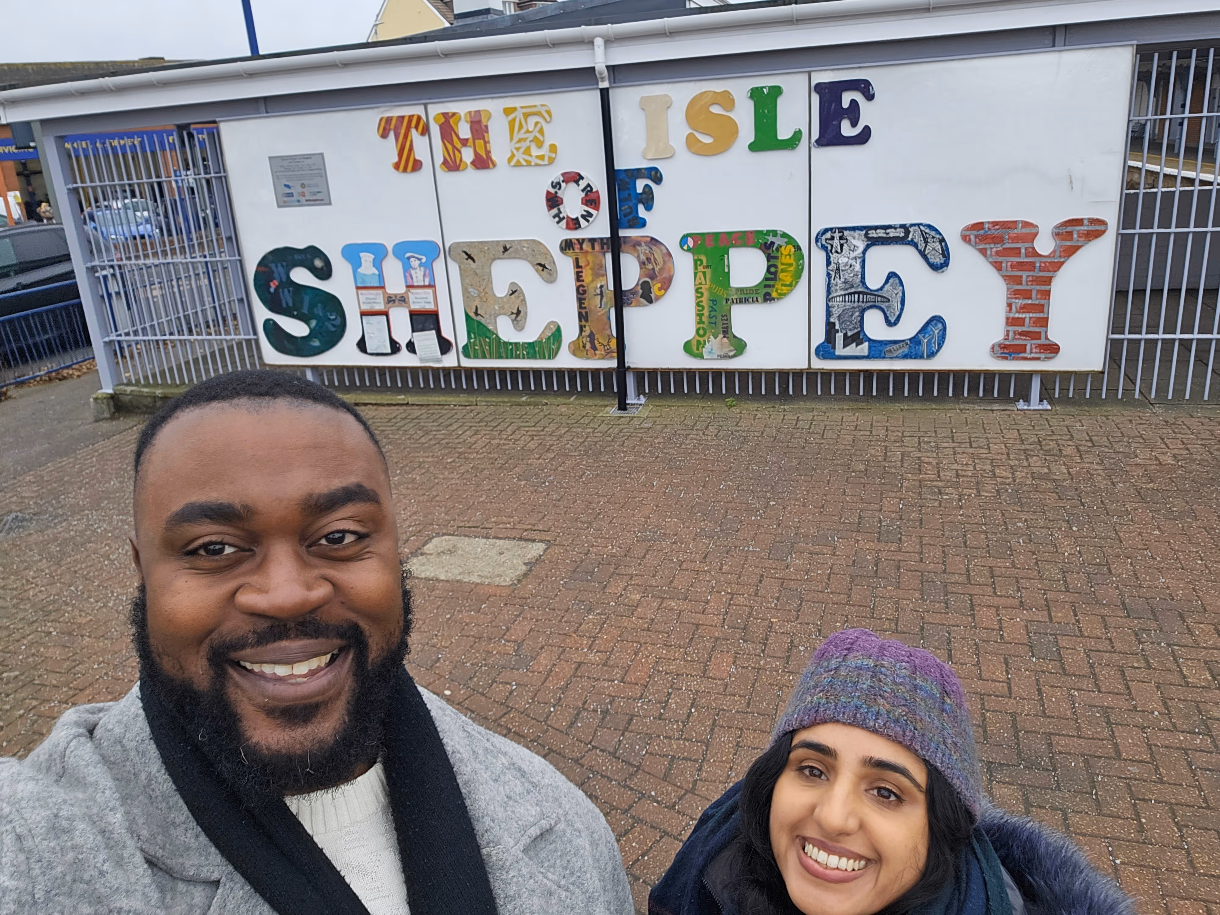 Smiling man and woman taking a selfie outdoors in front of a colorful sign that reads 'The Isle of Sheppey'.
