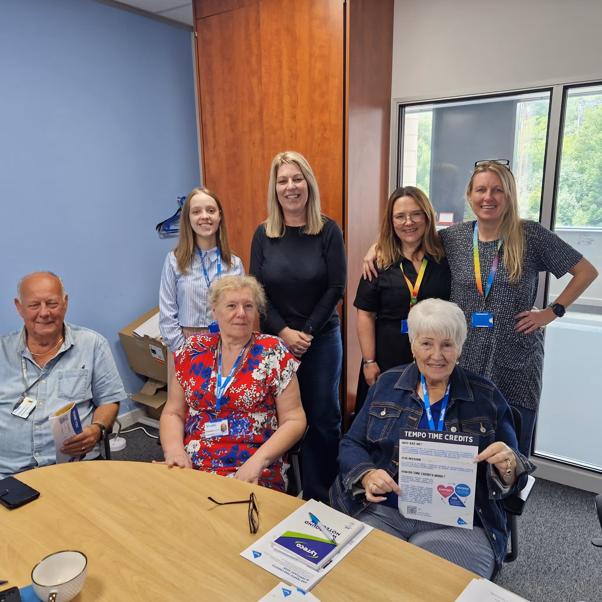 Group of seven people in an office, including elderly individuals and staff wearing lanyards, some holding informational pamphlets about Tempo Time Credits.
