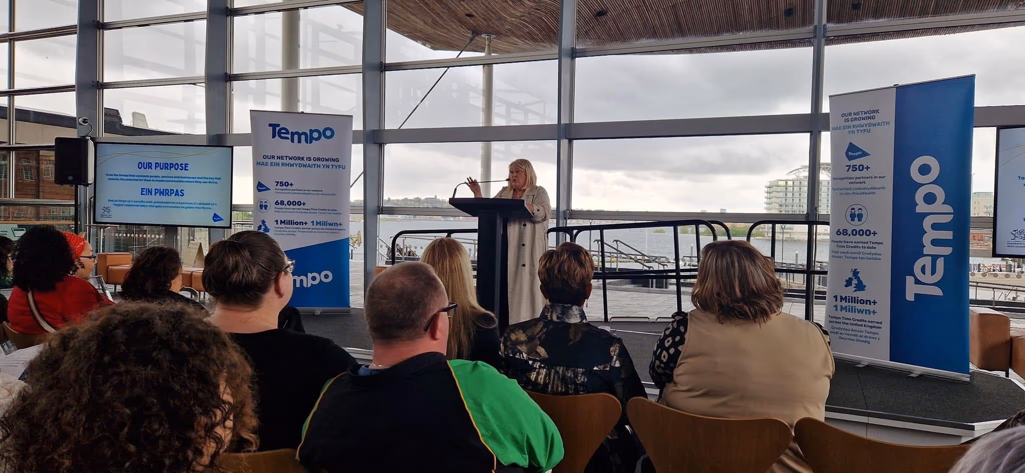Speaker at podium addressing an audience in a modern venue with Tempo banners and a view of a waterfront.
