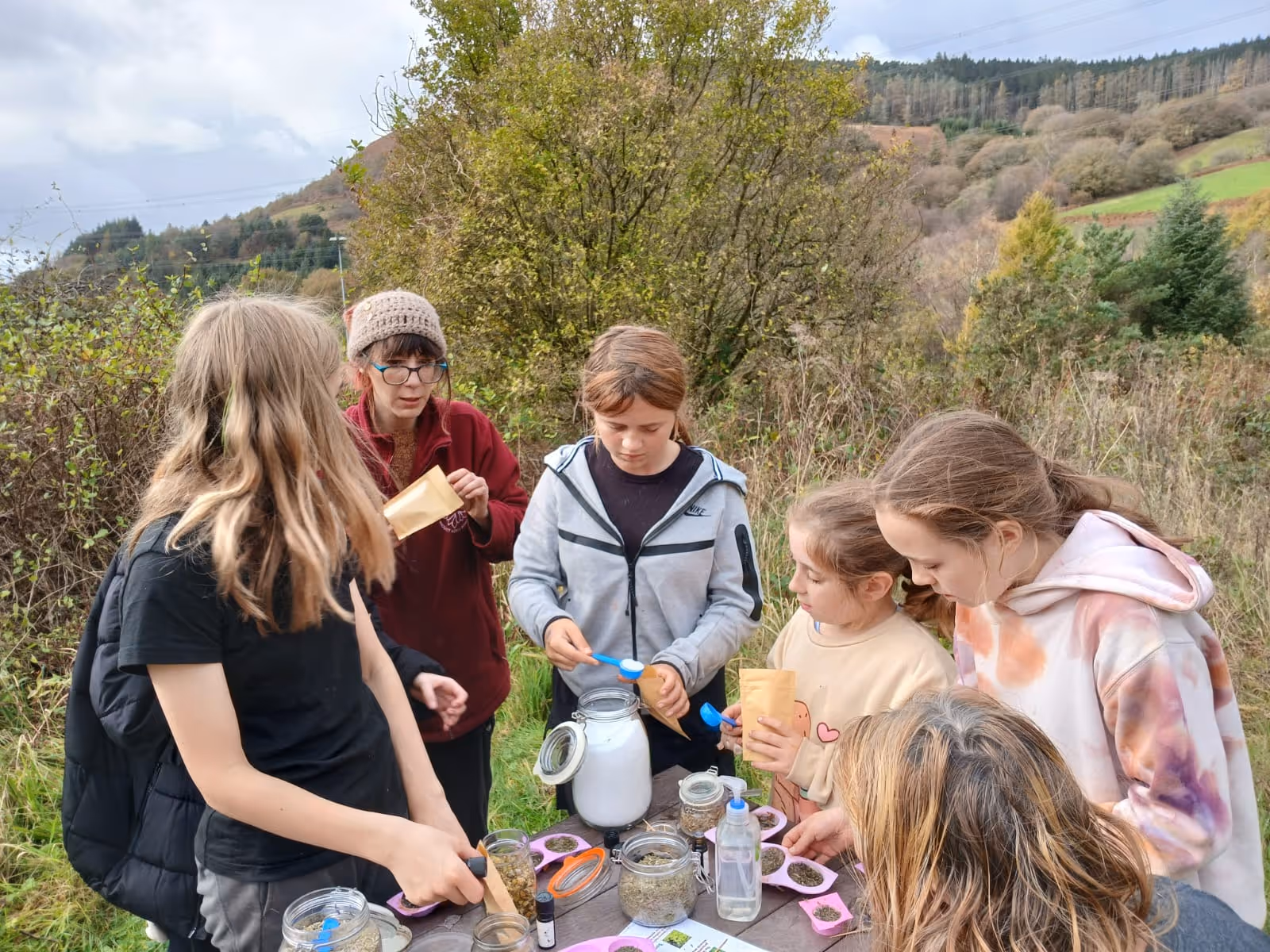Group of children and an adult outdoors gathering and measuring herbs or seeds at a wooden table with jars and scoops.
