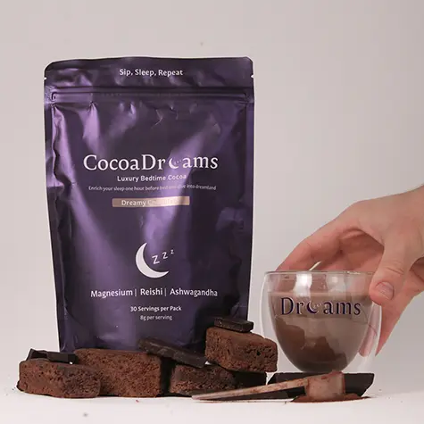 Hand holding a glass cup labeled Dreams next to a purple bag of CocoaDreams luxury bedtime cocoa with chocolate pieces in front.