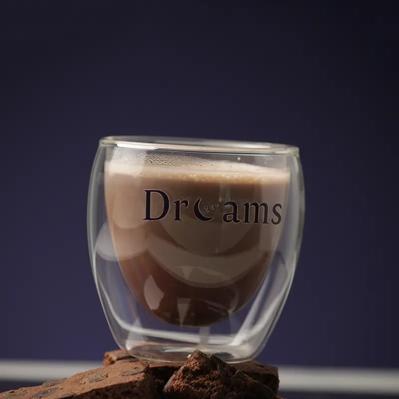 Double-walled glass cup with chocolate drink labeled 'Dreams' resting on chocolate cookies against a dark background.