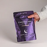 Hand holding a purple resealable pouch of CocoaDreams night cocoa powder with moon illustration.