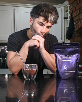 Man with dark hair and beard sitting at a kitchen counter with a glass of cocoa and a purple CocoaDreams package.