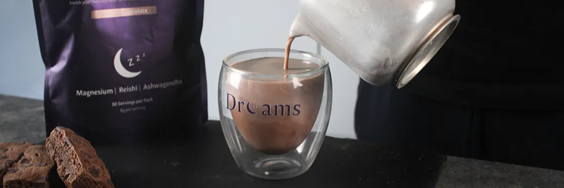 Hot chocolate being poured from a pot into a clear glass mug labeled 'Dreams' on a dark surface with chocolate brownies and a supplement bag in the background.