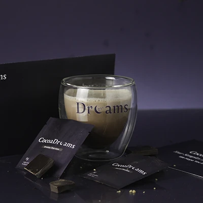 Glass cup with beige beverage labeled 'Dreams' surrounded by CocoaDreams branded dark chocolate pieces and packets.