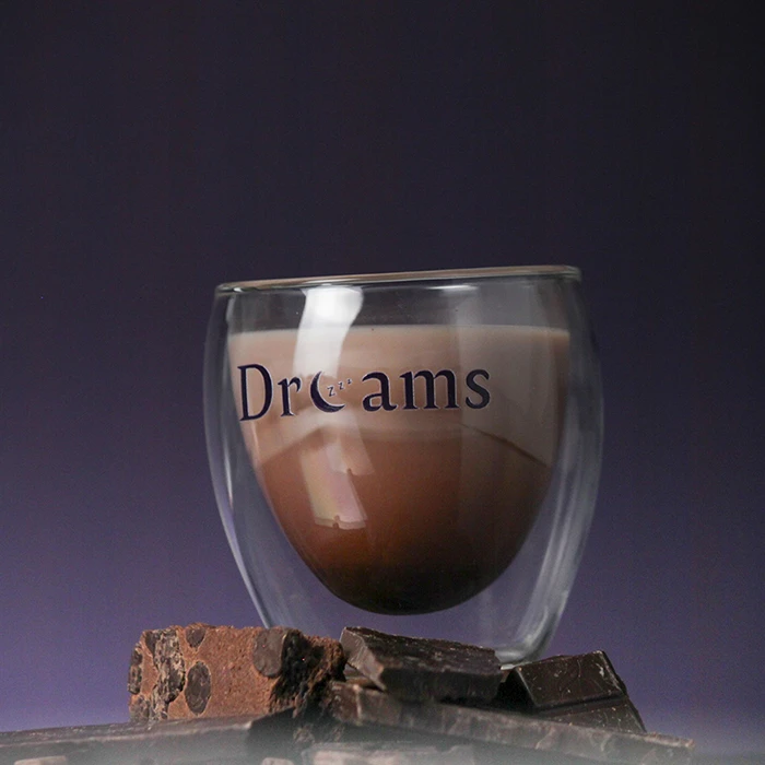 Double-walled glass cup filled with chocolate drink labeled 'Dreams' with pieces of chocolate and brownie in front.