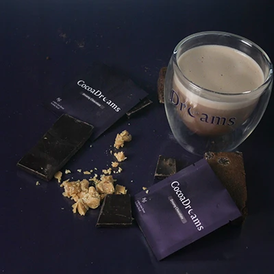 Glass cup with creamy beverage labeled 'Dreams' next to dark chocolate pieces and purple CocoaDrCams wrappers on a dark surface.