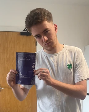 Young man wearing a white t-shirt with a green four-leaf clover holding a purple product package indoors.