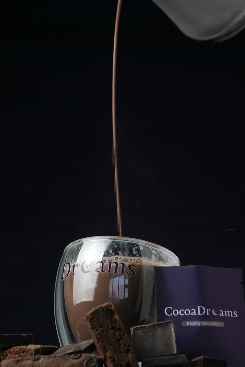 Chocolate drink being poured into a glass cup labeled 'Dreams' with scattered chocolate pieces and a CocoaDreams box nearby.