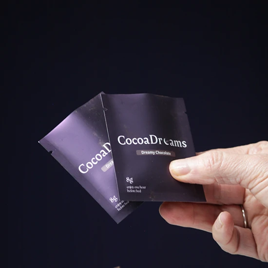 Hand holding two black packets of CocoaDreams Dreamy Chocolate powder against a dark background.