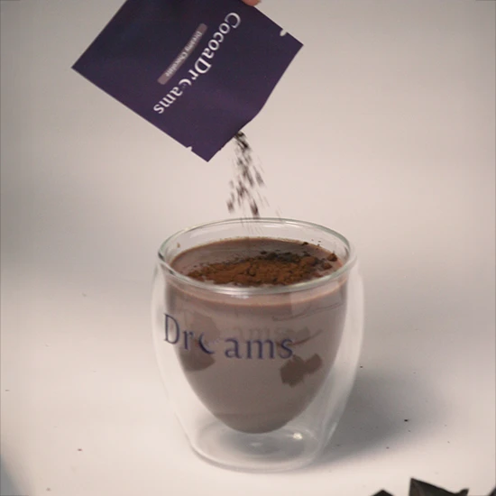 Cocoa powder being poured from a packet labeled 'CocoaDrams' into a double-walled glass cup with the word 'Dreams' printed on it.