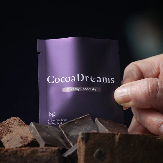 Hand holding a purple CocoaDreams packet labeled Dreamy Chocolate above chunks of dark chocolate and cocoa powder.