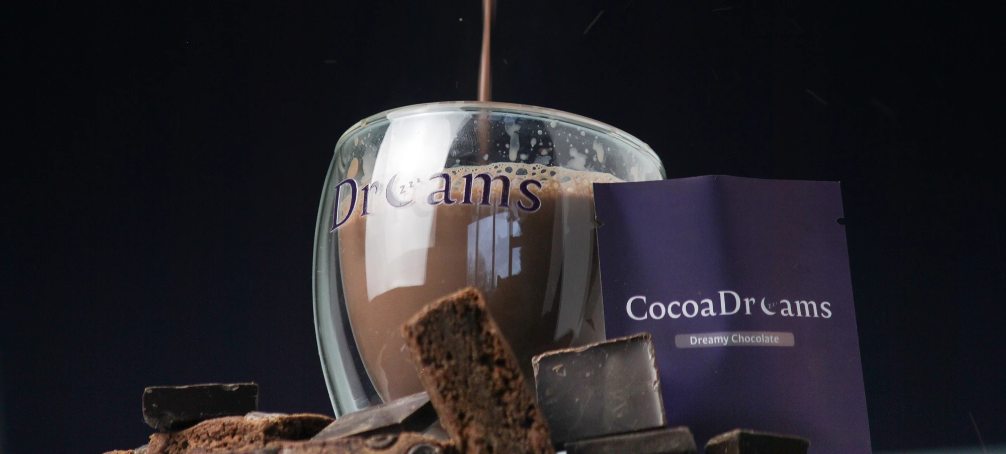 Glass mug labeled Dreams being filled with hot chocolate surrounded by chocolate pieces and CocoaDreams packaging.