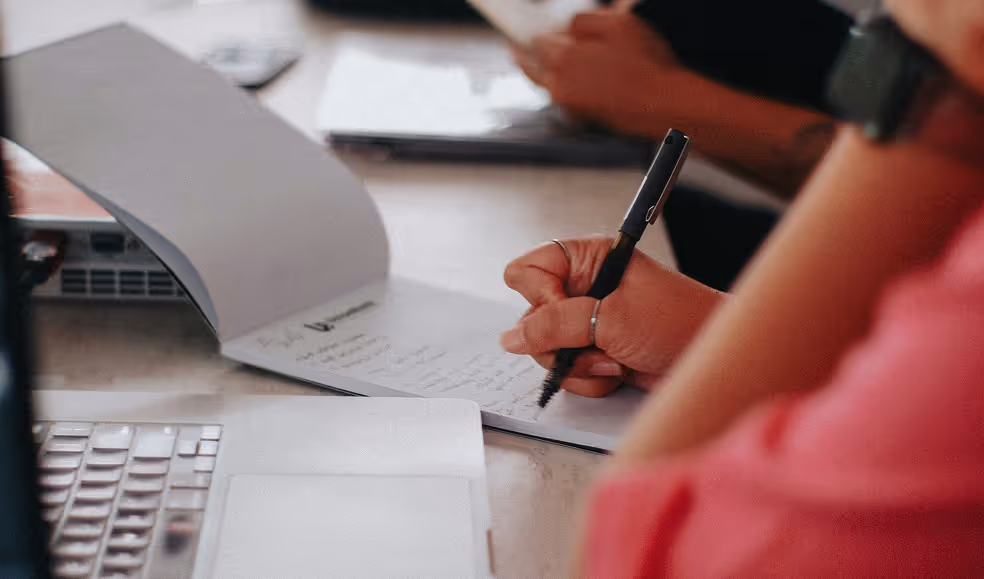 A person writing notes with a pen on a notepad next to a laptop and another person in the background.