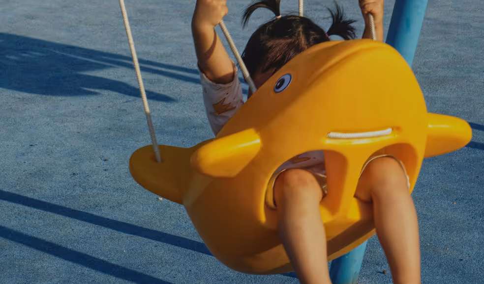 Child with pigtails swinging on a yellow fish-shaped playground swing.