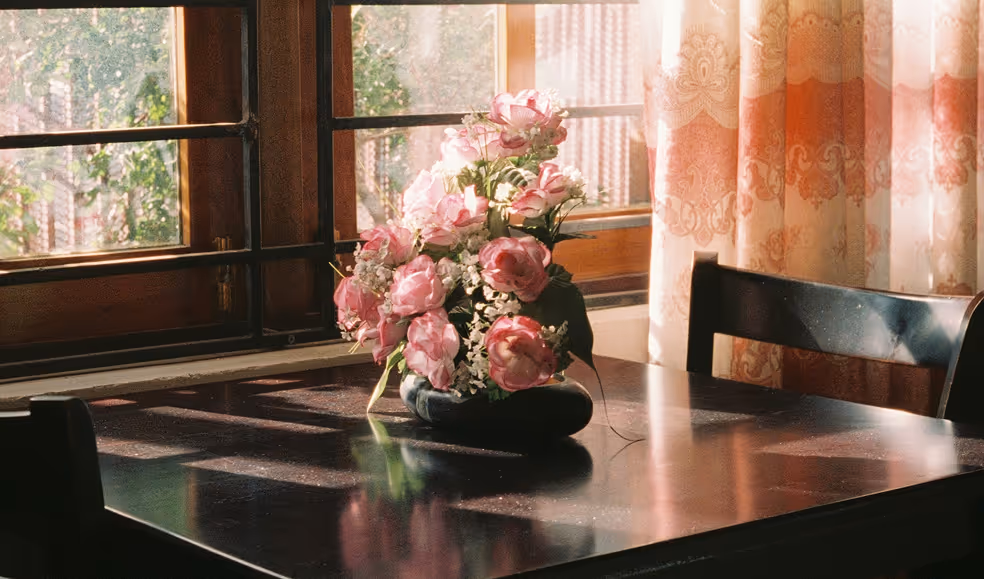 Bouquet of pink peonies in a vase on a wooden table near sunlit window with floral curtains.