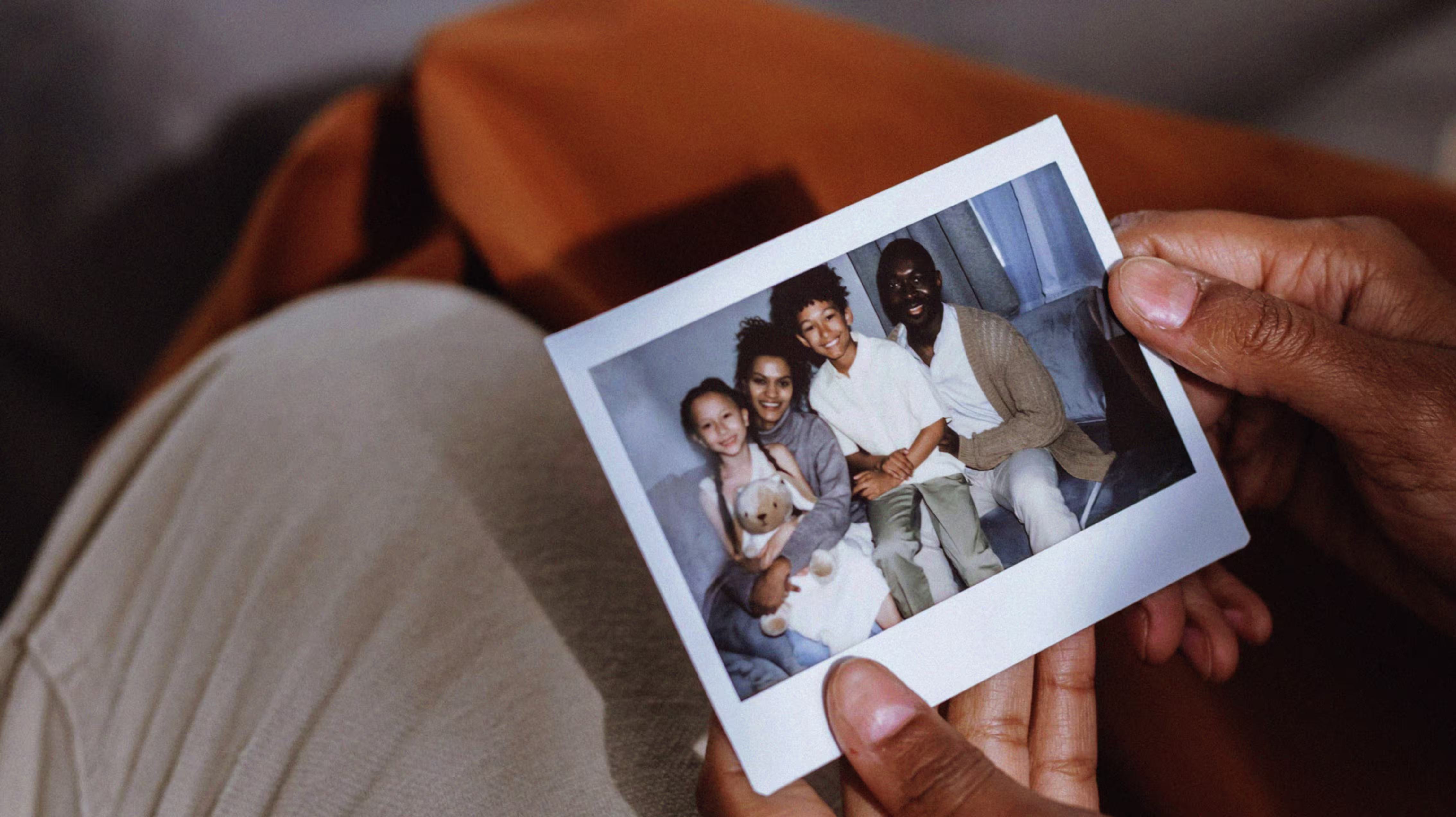 Hands holding a printed photo of a smiling family of four sitting together on a couch.