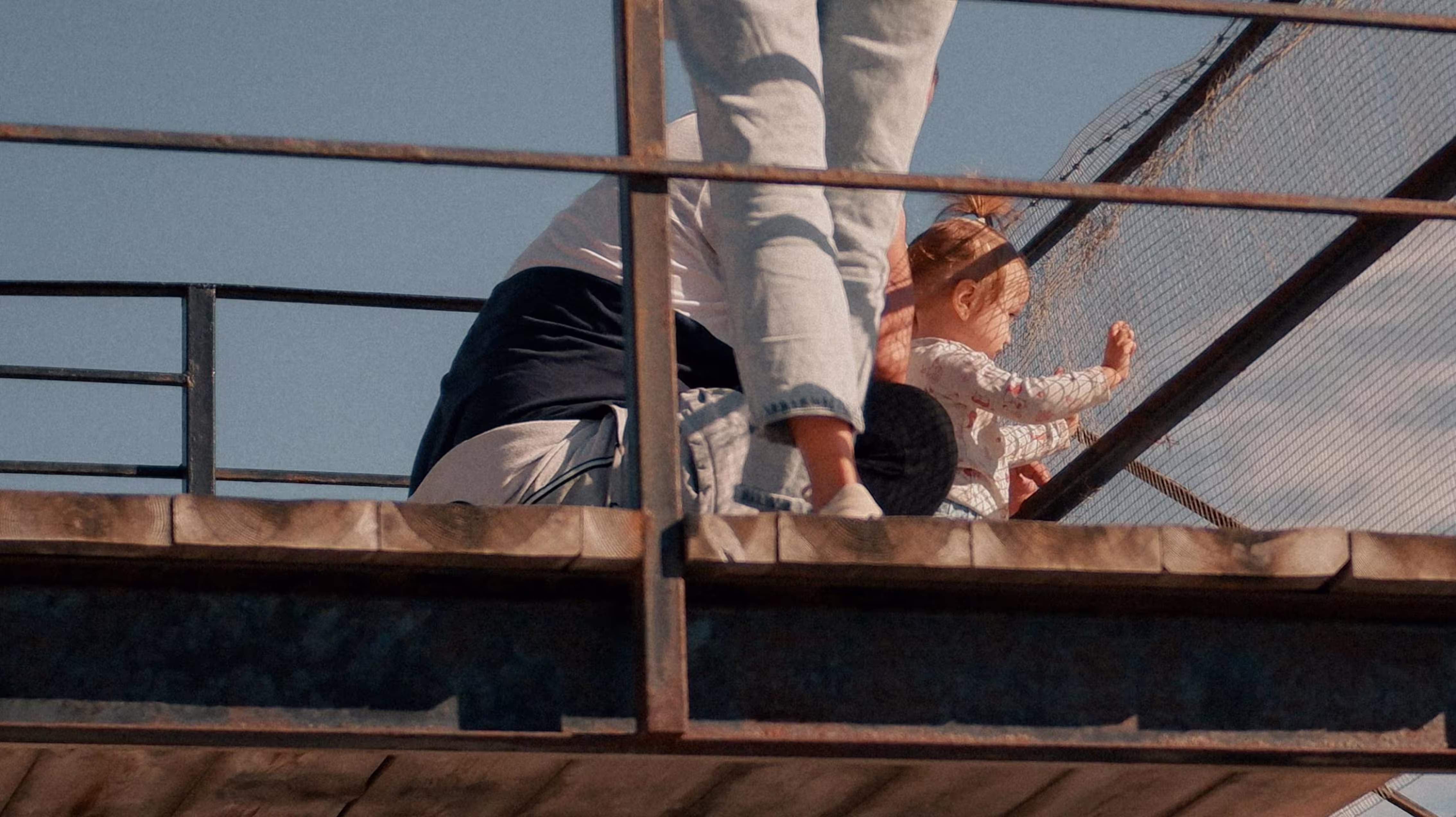 Child and adult sitting on a wooden platform behind a metal railing and wire fence against a clear sky.