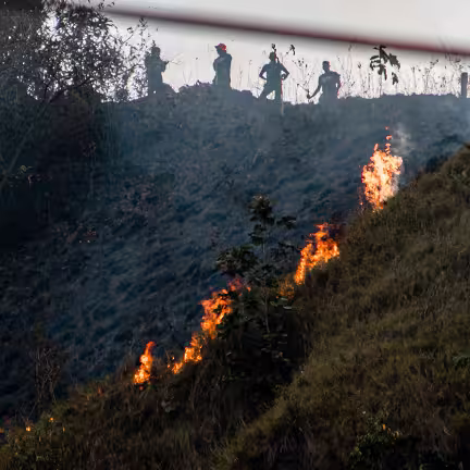 Wildfire burning on a hillside with silhouettes of firefighters standing at the ridge above.