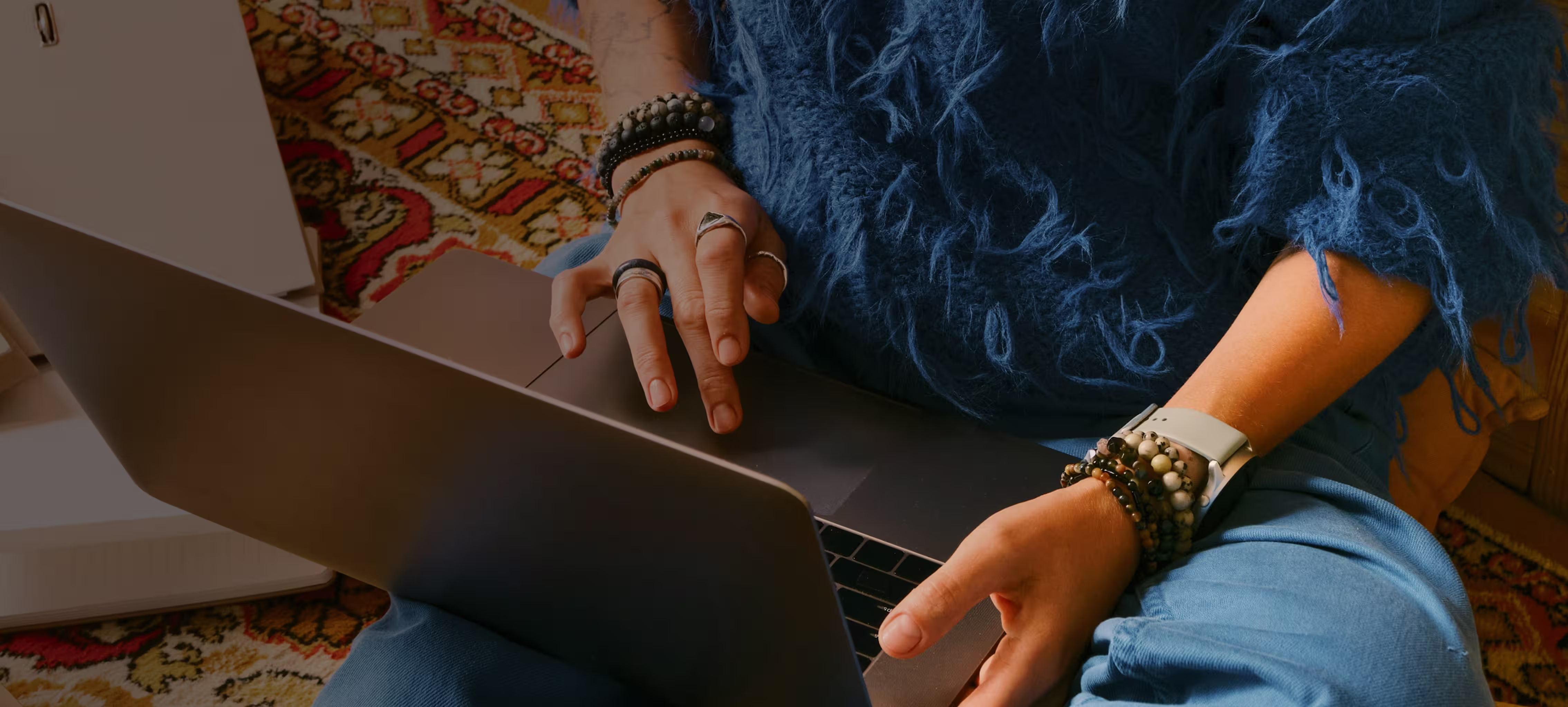Person sitting cross-legged on a colorful patterned carpet, typing on a laptop while wearing a blue fuzzy sweater and multiple bracelets.