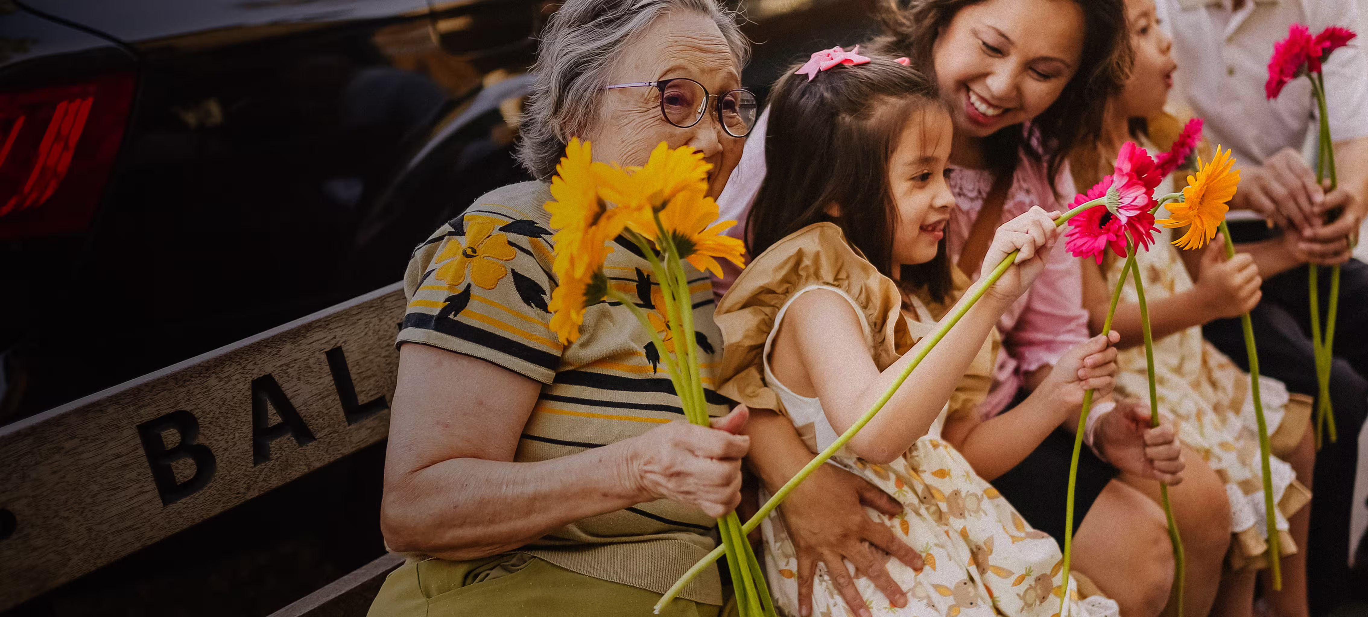 Smiling multi-generational family sitting on a bench holding colorful flowers.